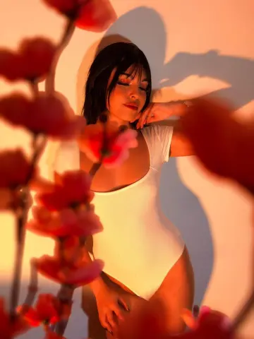 DapneOwen poses in a white bodysuit with red flowers in the foreground and a warm shadow behind her.