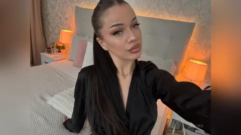 AriaVex poses in a black silk blouse against a light blue headboard and warm bedside lamps.