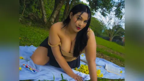 Curvy woman in a tan top sitting on a white picnic blanket in a lush green park setting.