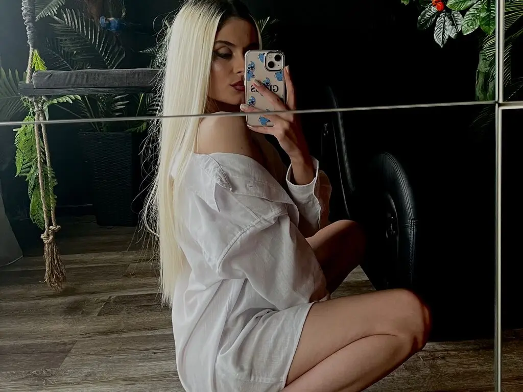 Blonde woman in a white oversized shirt taking a mirror selfie in a moody, plant-filled room.