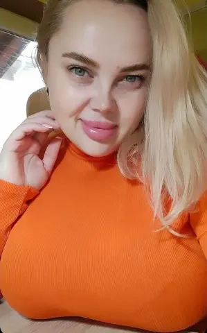 Smiling blonde woman in a bright orange ribbed turtleneck sweater posing indoors.