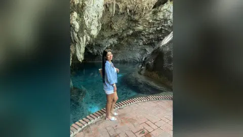 Young woman in a blue shirt standing by a turquoise cave lagoon under rocky stalactites in a serene setting.