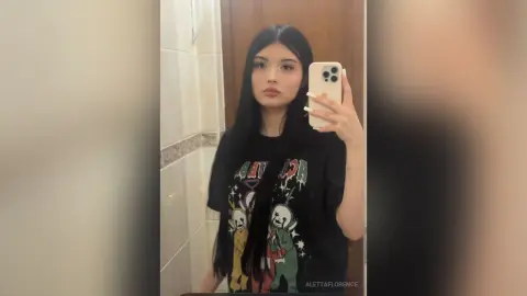 AlettaFlorence poses for a mirror selfie wearing a black graphic t-shirt, showing off her long, straight black hair.
