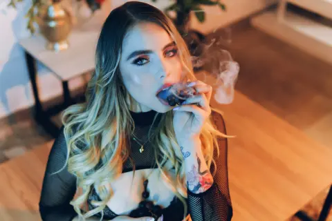 Blonde woman in a black mesh top smoking a cigar in a cozy, dimly lit indoor setting.