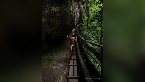 ElizabethSwith stands naked on a wooden walkway in a lush, green forest, posing with her hands up near her head against a large rock wall.