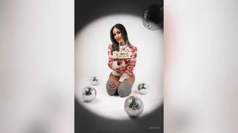 Glamorous woman in a festive red and white sweater posing with disco balls in a bright studio setting.