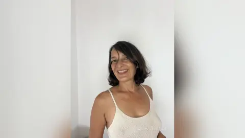 MichaelaBelle smiles while wearing a white ribbed tank top against a plain white wall.
