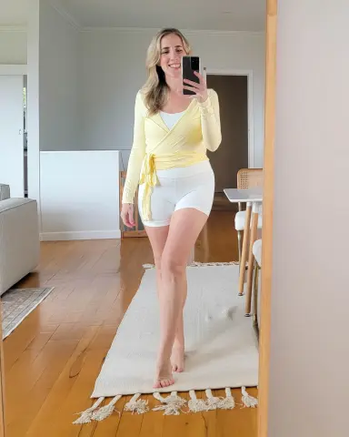 Smiling woman in a yellow wrap top and white shorts taking a mirror selfie in a bright living room.