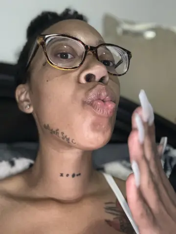 LexBright pouts her lips while wearing clear-lens glasses and showing off her long white nails and neck tattoo.