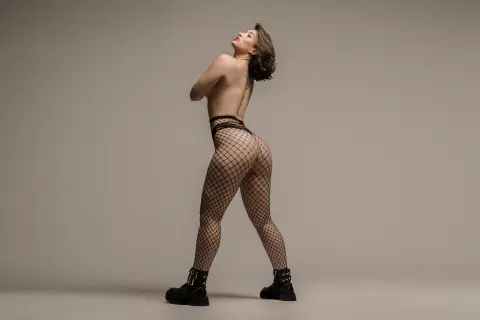 Katt_Henao poses naked from the waist up, wearing black fishnet tights and black boots against a plain grey background.