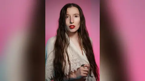 Young woman with long wavy hair wearing a white lace blouse against a soft pink studio background.