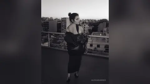 Moody black and white portrait of a woman in a black outfit standing on a city rooftop at dusk.