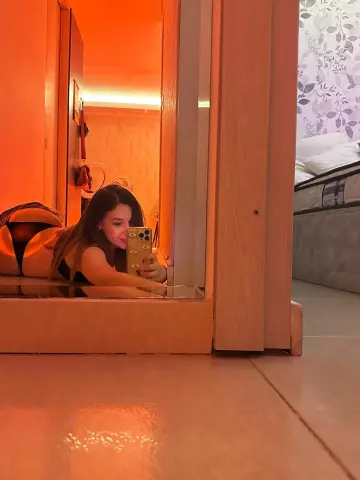 Young woman in black outfit taking a mirror selfie in a warm, cozy bedroom setting.