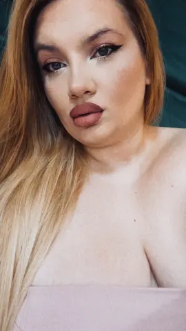 Close-up portrait of a blonde woman with winged eyeliner and mauve lipstick posing in a soft, natural light.
