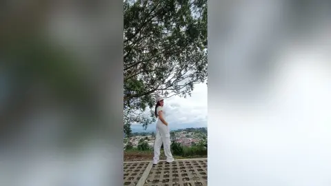 AmelieLautrec stands outdoors in a white sleeveless top and white pants, posing next to a large leafy tree overlooking a town view.
