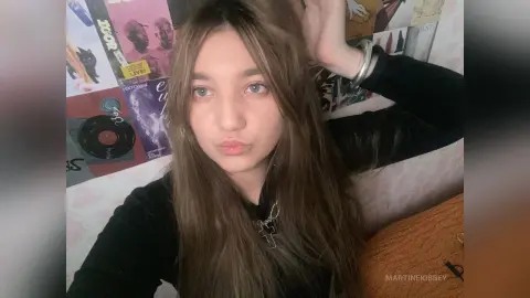 MartineKibbey poses with long brown hair and a black long-sleeve top, resting her hand behind her head against a wall of music posters.