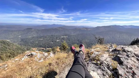 Relaxed hiker in black leggings resting on a rocky mountain peak overlooking a vast green forest valley.
