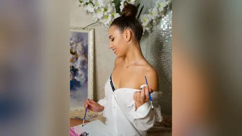 DelanaGrace smiles while wearing an open white button-down shirt and a black bra, holding a blue paintbrush near a canvas.