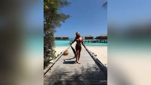 Slim woman in a brown bikini walking on a wooden pier at a tropical beach resort under a clear blue sky.