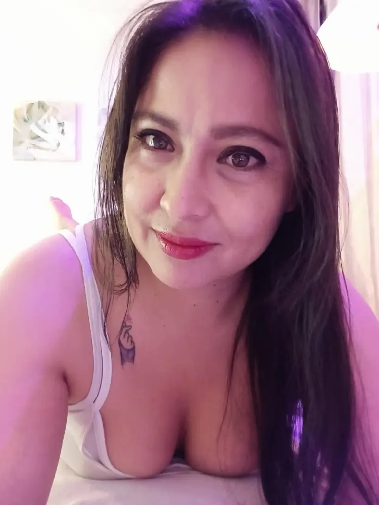 Hanna_backerr lies on a bed wearing a low-cut black top that shows off her cleavage and dark hair.