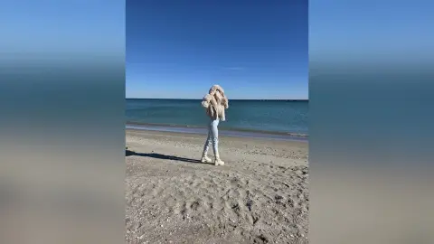 Young woman in a cozy beige sweater and jeans standing on a sunny beach by the blue ocean.