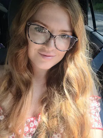 Anna_OG smiling in glasses with long, wavy red hair, wearing a red and white floral top while sitting in a car.