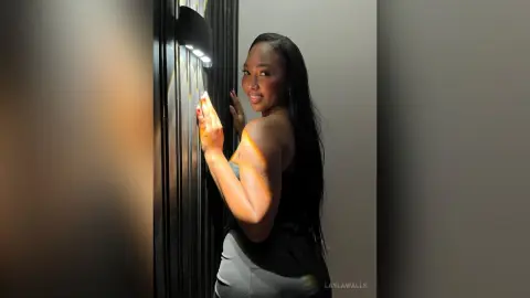 Smiling Black woman in a black dress posing by a wall sconce in warm, moody indoor lighting.