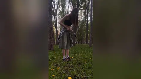 Young woman in a black tank top and olive skirt standing in a lush green forest setting.