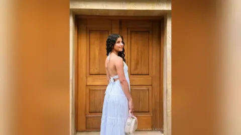 Elegant woman in a white backless summer dress standing before a rustic wooden door in soft natural light.