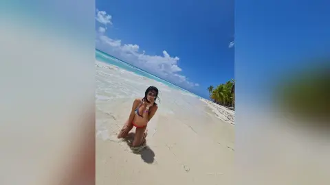 SandraMidnight poses naked or in red bikini bottoms while kneeling on a white sand beach under a blue sky.