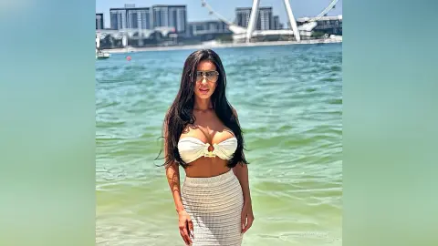 Brunette woman in a white bikini top and ribbed skirt standing by the ocean under bright sunlight.