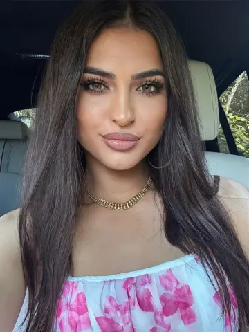 Young woman with long dark hair wearing a floral white top and gold chain necklace in a car seat.