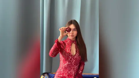 Young woman in a red lace dress holding a pomegranate in front of her eye against a blue curtain background.