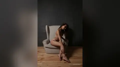 LeahEvans sits on a grey armchair in light lingerie, posing with her legs crossed against a dark wall.