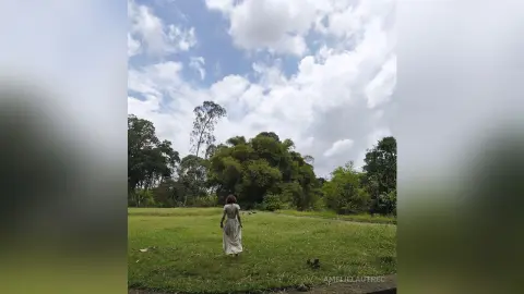 AmelieLautrec stands in a grassy field wearing a long white dress, facing away from the camera toward a dense line of green trees under a cloudy sky.