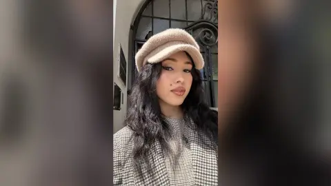 Young woman with long dark hair wearing a beige cap and houndstooth sweater in a bright outdoor setting.