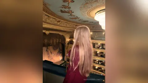Blonde woman in a burgundy outfit admiring the ornate gold ceiling of a luxury classical opera house.