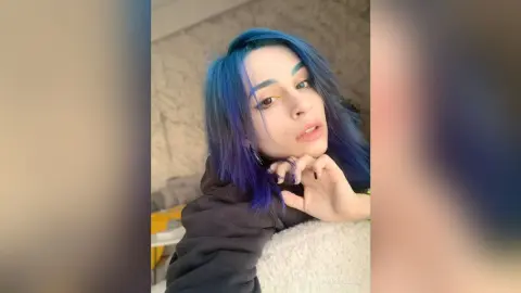Young woman with vibrant blue hair and casual black sweatshirt smiling in a cozy indoor setting.