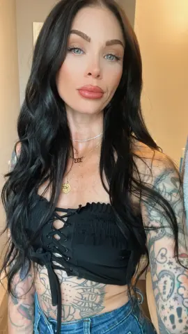 Stacy_babyy poses with long black hair, wearing a black lace-up crop top and blue jeans, showing off her tattoos on her arms and stomach.