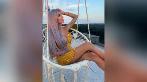 Woman with long lavender hair in a yellow polka dot swimsuit relaxing on a white hanging chair at sunset.