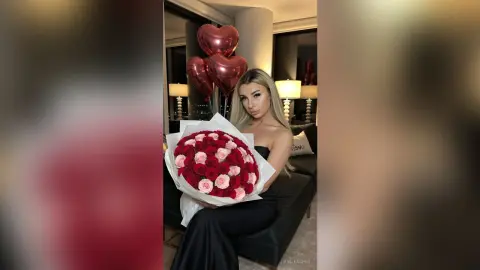 Elegant woman in a black dress holding a large bouquet of red roses with heart balloons in a cozy room.