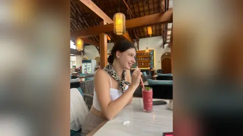 RitaMoonllight sits at a table in a restaurant, wearing a white tank top and a leopard print scarf, holding a pink drink with a straw.