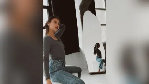 Stylish woman in a grey long-sleeve shirt and blue jeans posing in a minimalist, bright studio setting.