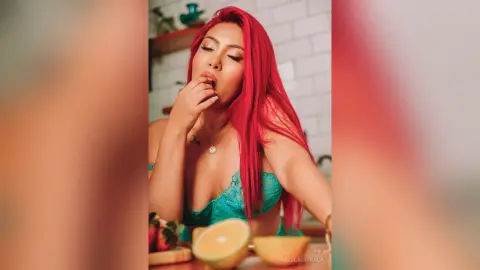 IsabellaEstrada licks her finger near her mouth, wearing a teal lace bra with long red hair in a kitchen setting.