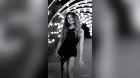 Smiling woman in a black cocktail dress posing under glowing outdoor lights in a moody black and white portrait.