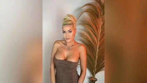 Glamorous blonde woman in a brown strapless dress posing elegantly against a tropical palm leaf backdrop.