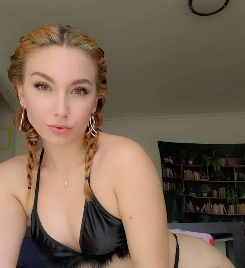 Rebelrebbb poses with her long blonde hair, wearing a black choker and a black top against a dark background.