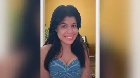 Smiling woman in a blue denim corset top relaxing in a bright, sunlit indoor setting.