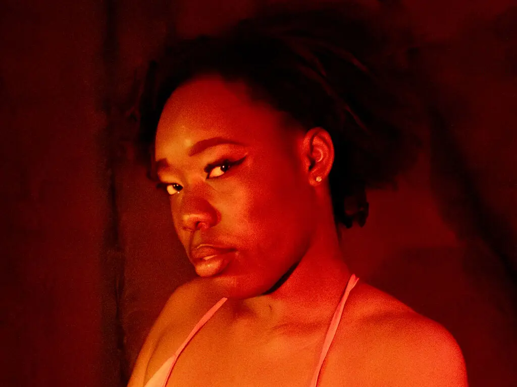 Moody portrait of a Black woman in a white strap top under warm red lighting.
