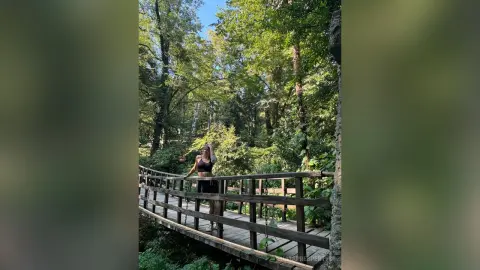 SilviaPrespertys poses in a black crop top and shorts on a wooden walkway surrounded by lush green trees.